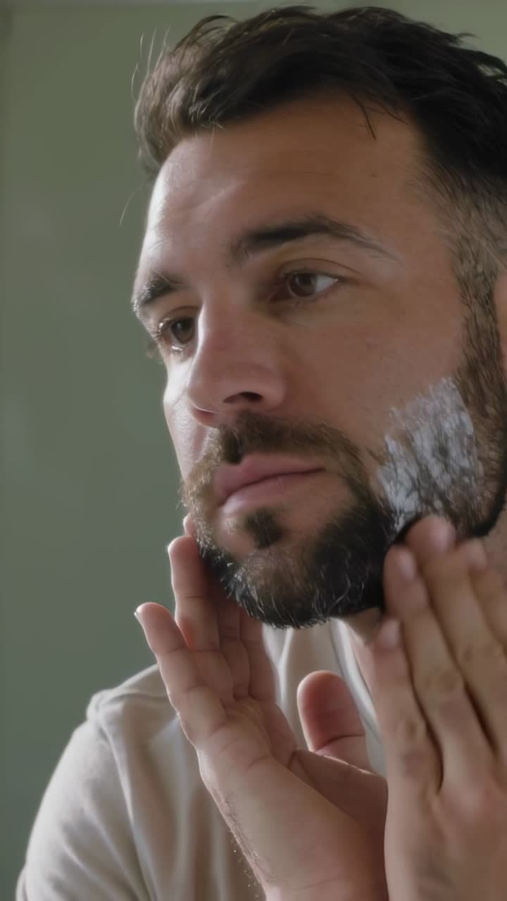 Video verticale: Raccogliere schiuma, uomo che la applica sulla guancia sinistra in bagno indossando una maglietta, controllando la barba