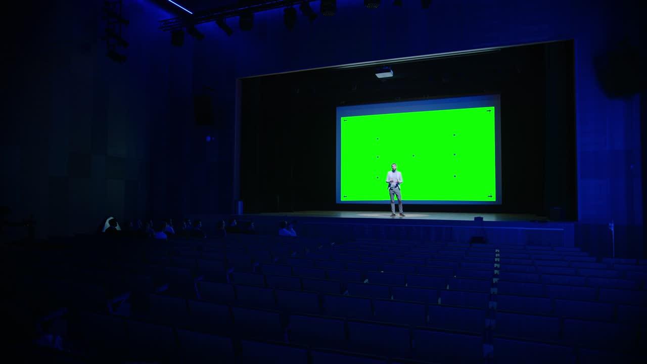 Keynote Speaker Does Presentation of New Product to the Audience, Behind Him Movie Theater with Green Screen, Mock-up, Chroma Key. Business Conference Live Event or Device Reveal