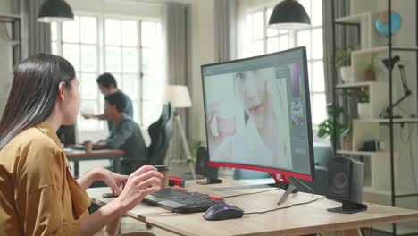 asian female digital editor using mobile phone while works in photo editing software on his personal computer in office