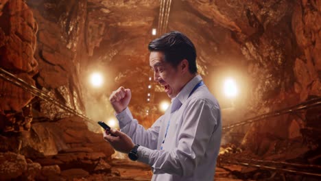 side view of an asian male professional worker with his smartphone in underground mine tunnel, he raises his fist up with screaming goal after check on the smartphone