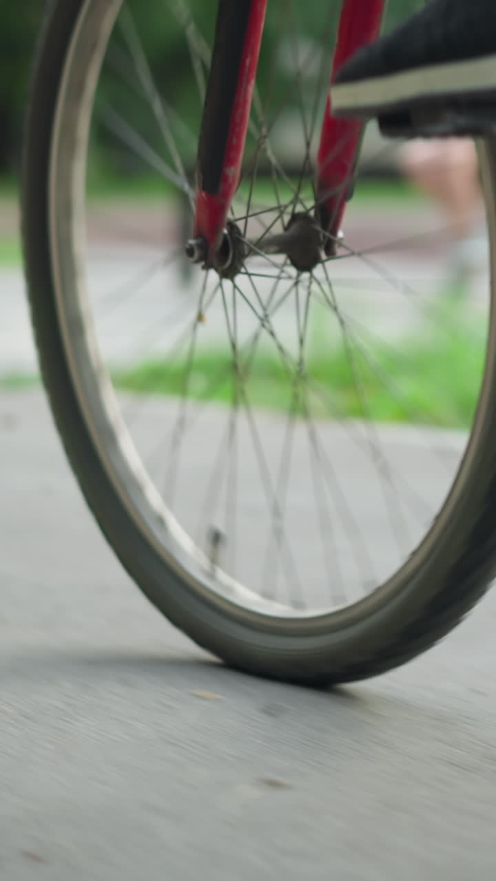 vista de la pierna de alguien con zapatillas de deporte montando una bicicleta en una carretera pavimentada, capturada desde un ángulo bajo, en el fondo, una figura borrosa está sentada en un banco cerca de la vegetación y los árboles