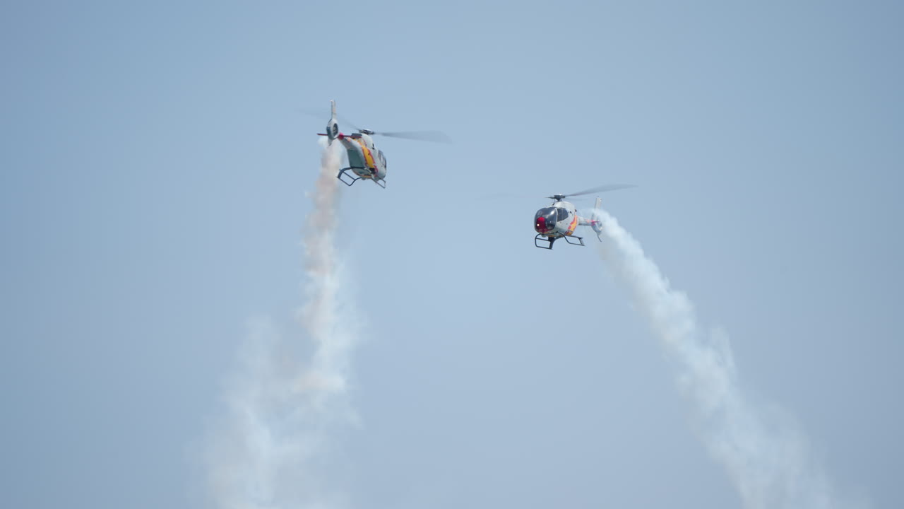 Two helicopters performing synchronized aerial stunt with smoke trails in clear blue sky