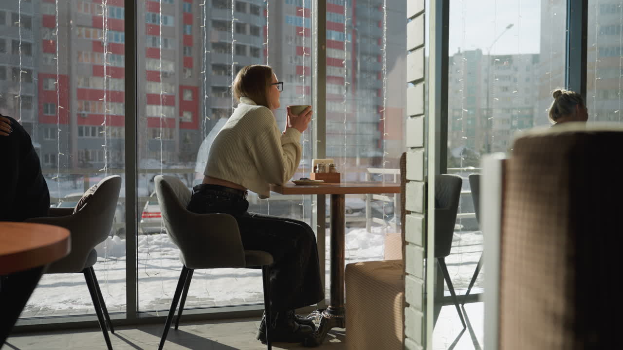 Side view of young woman in glasses sipping coffee inside eatery during winter, sitting near window with urban residential buildings outside, daylight casting warm shadows, other patrons nearby