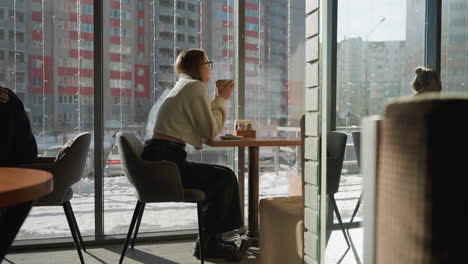 side view of young woman in glasses sipping coffee inside eatery during winter, sitting near window with urban residential buildings outside, daylight casting warm shadows, other patrons nearby