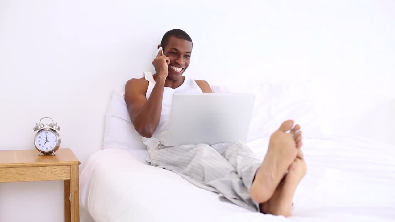 Handsome man lying on bed phoning and using laptop