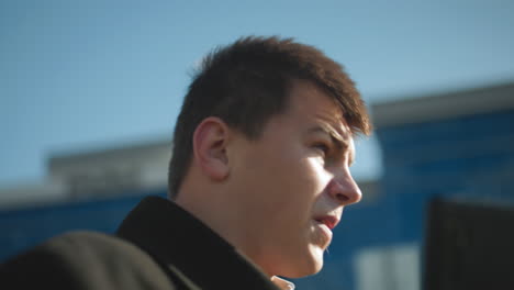 close-up man in black coat on phone call outdoors, shaking head in displeasure while reacting to intense conversation, urban background with modern buildings, greenery, and clear blue sky