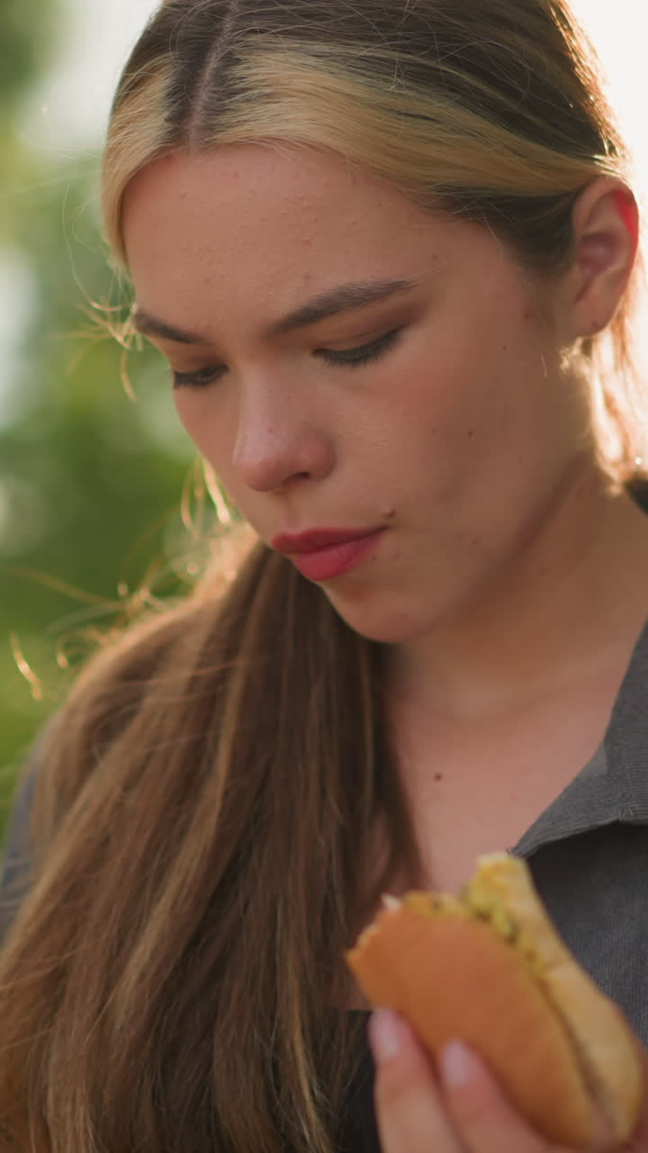 primer plano de una mujer con una camiseta gris sosteniendo shawarma, observándola pensativamente, la luz del sol suave ilumina su mano y el bocadillo, con un fondo borroso de vegetación y cálidos rayos de sol