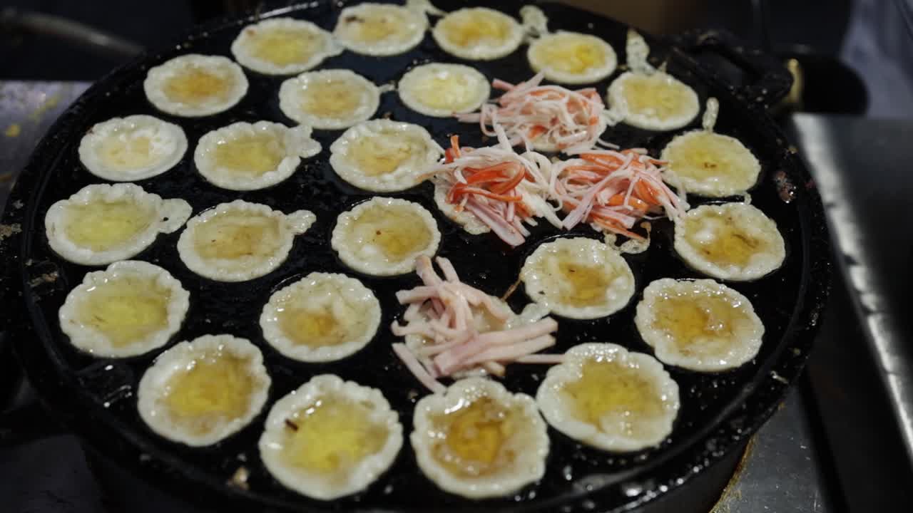 vendedor de alimentos poniendo jamón en mini panqueques fritos crujientes usando pinzas en una sartén de takoyaki de estilo tailandés en el mercado