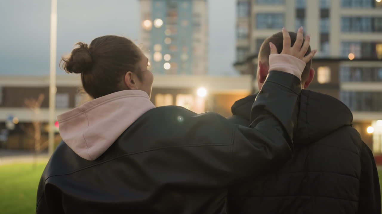 Back view of woman playfully touching friend head while strolling together, light from residential building in background creating a warm and relaxed evening vibe