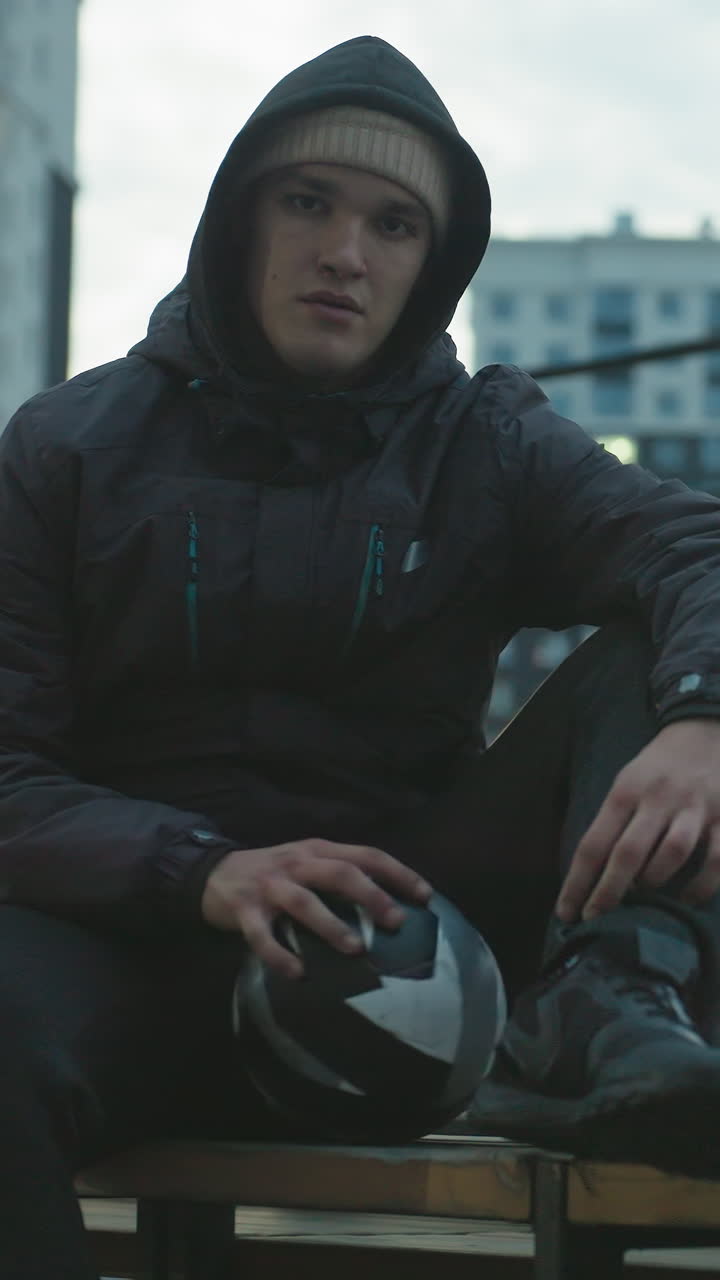 hombre con capucha sentado en un banco girando una pelota de fútbol durante una sesión de entrenamiento al aire libre en una arena deportiva urbana, rodeado de modernos edificios residenciales con luces brillantes de la noche