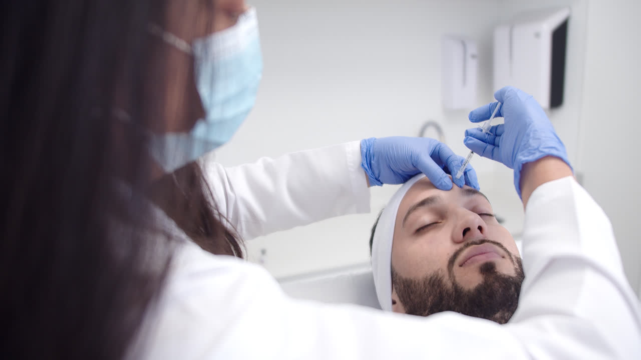 Beautiful slow motion shot of a female doctor injecting a young Latino man in the face while he is lying on a table inside a dermatology clinic.
