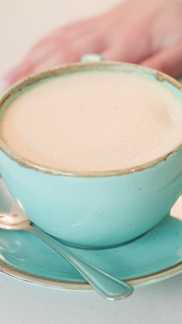Крупный план: руки белой женщины держат бирюзовую чашку, женщина держит латте в керамической чашке на подходящем блюдце с ложкой, теплый мягкий утренний свет, пастельные тона, нежная текстура пены, видимый маникюр, дом