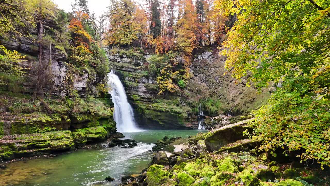 Cachoeira Saut du Doubs na França com água verde cristalina, pedras cobertas de musgo e uma vibrante floresta de outono. Penhascos íngremes e folhagens coloridas emolduram o rio que corre, criando uma paisagem natural tranquila.