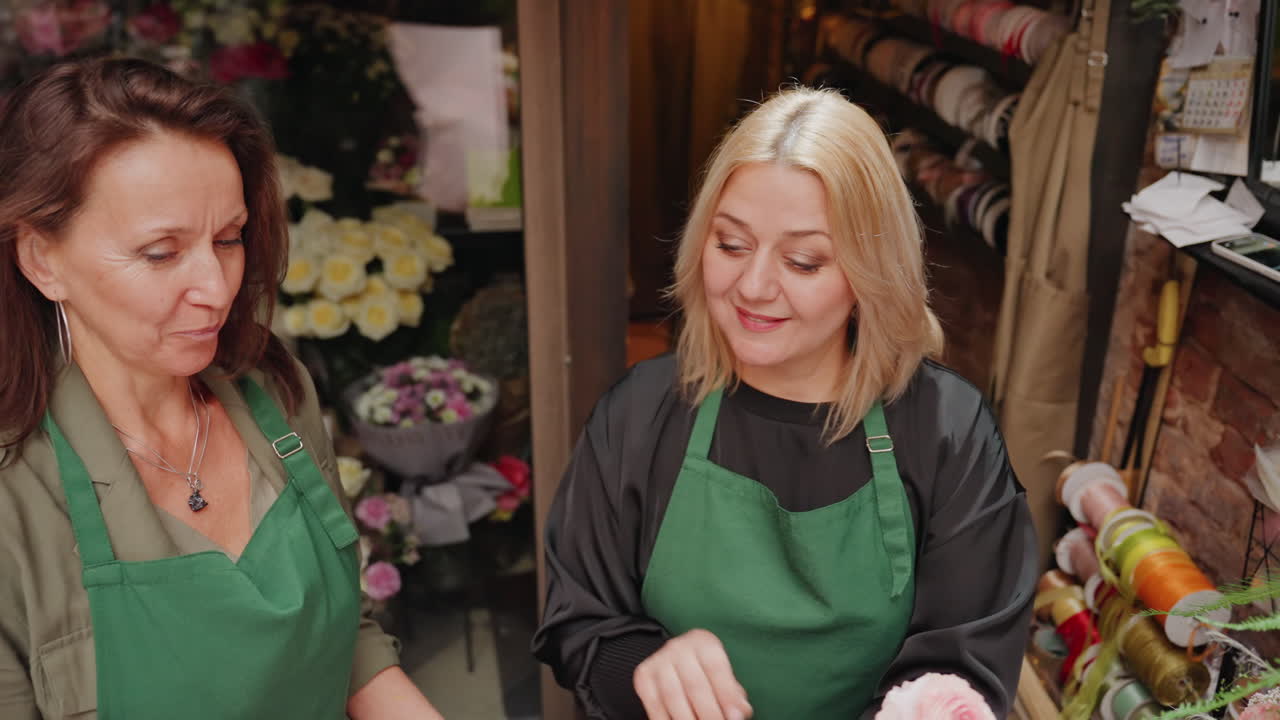 Zwei kaukasische Floristinnen wickeln gemeinsam einen rosa Blumenstrauß an der Theke ein. Kooperative Ladenszene mit Mentorin und Lehrling, die Papier und Band austauschen, grüne Schürzen, Schere und Bindfaden, Rosenstrauß.