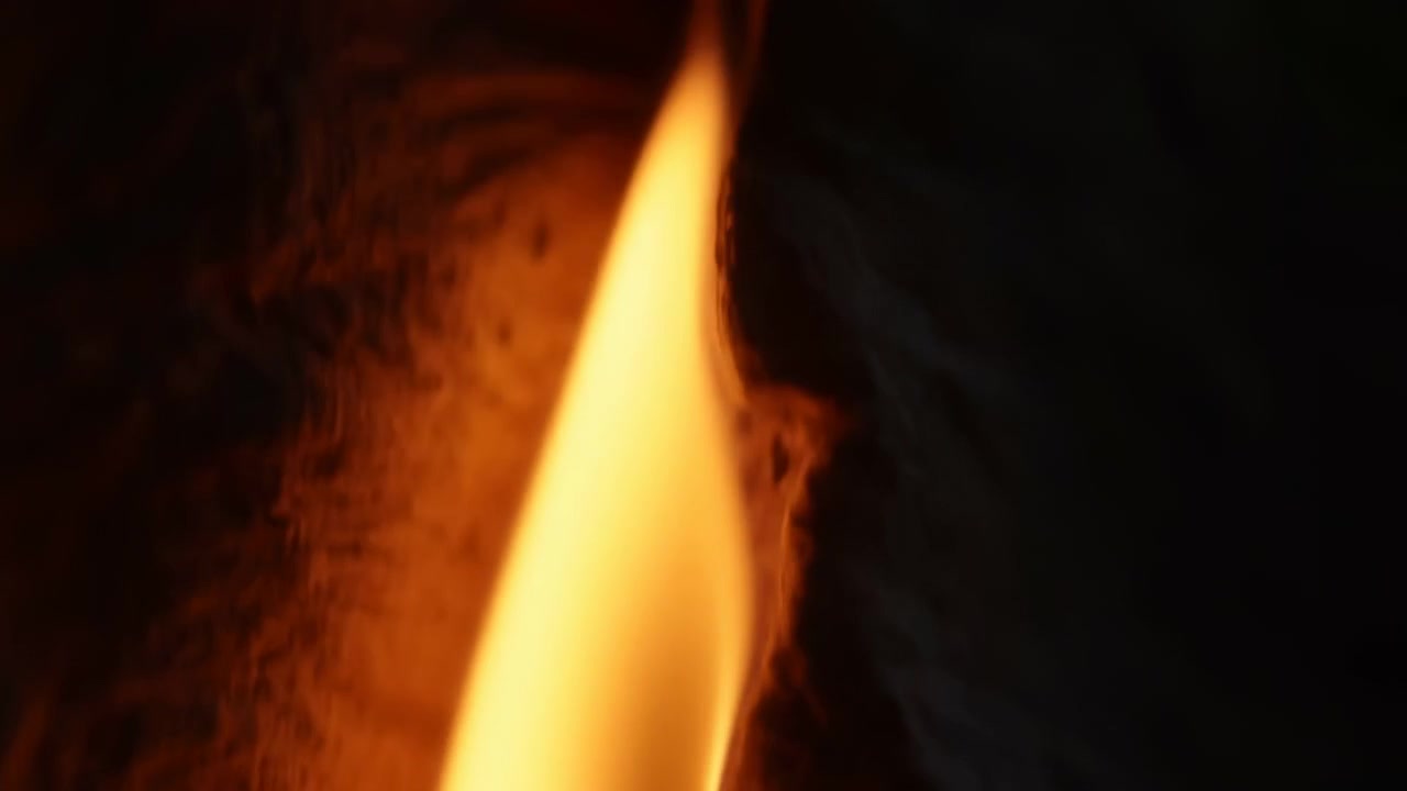 Tightly framed scene of narrow vertical flame or concentrated light column rising with slight rightward lean, bright yellow orange core, soft glow, and strong contrast over rough dark surface