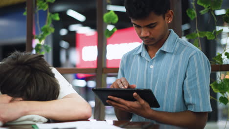 white boy resting head on sweater shoulder while asleep beside awake friend tapping tablet screen under subtle indoor light highlight contrast between restful slumber and focused digital interaction