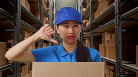 primer plano de una mensajera asiática insatisfecha en uniforme azul mostrando el pulgar hacia abajo mientras entrega una caja en el almacén