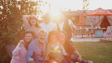 Portrait-Of-Mature-Friends-Sitting-Around-Fire-At-Outdoor-Campsite-Against-Flaring-Sun