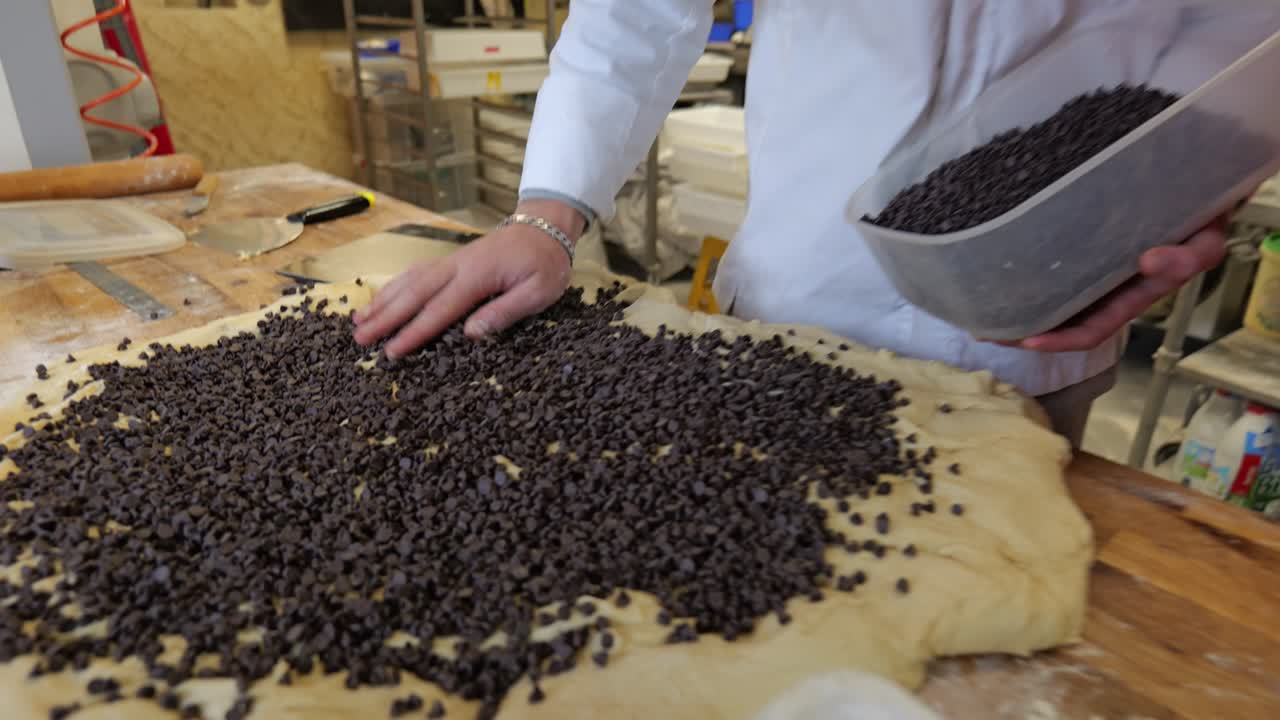 Primo piano di un pasticcere che sparge uniformemente gocce di cioccolato su impasto crudo per dolci, a mano, su una superficie di lavoro in legno all'interno di una pasticceria tradizionale.