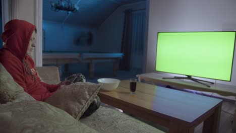 a man sits in the evening on a couch with a mobile phone in front of a green screen tv