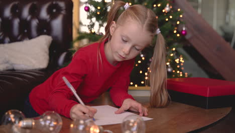 blonde girl sitting at the table writing on the paper near the christmas tree