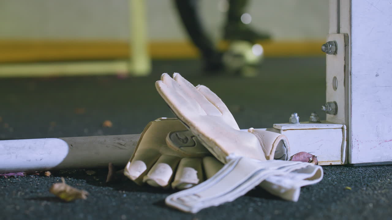 색 골키퍼 장갑이 골포스트 근처에 잔디에 어져있는 건조한 잎, 밤에 도시 조명에 의해 조명 된 발 에서 공을 균형 잡는 축구 선수의 흐릿한 배경