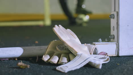 primer plano de guantes blancos de portero descansando cerca del poste de la portería en el césped con hojas secas esparcidas, fondo borroso de un jugador de fútbol equilibrando la pelota bajo los pies iluminado por las luces urbanas por la noche