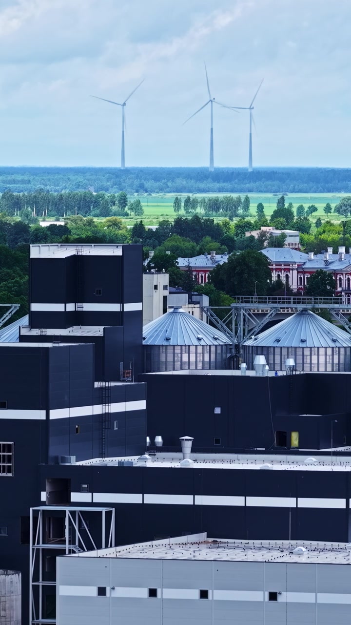 Modern industrial plant complex and silos with rotating wind turbines in the distant green fields