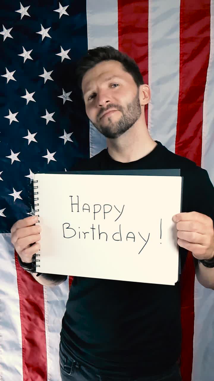 Tattooed man lifts up a notepad that has Happy Birthday written on white paper, while the man is standing in front of a waving American US flag. Celebrating independece day or 4th of July with a smile