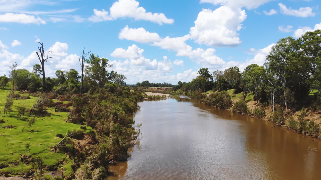 volando río abajo de un río marrón, sucio y turbio con hermosos bancos verdes cubiertos de hierba a cada lado mientras que las nubes blancas y esponjosas flotan arriba en el pintoresco cielo azul