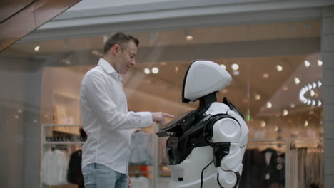 a man stands with a robot bot and asks him questions and asks for help by clicking on the screen on the robot body. human-robot interaction in the modern world