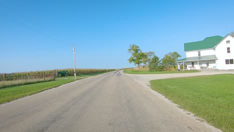 POV-driving-on-rural-county-road-past-maturing-field,-farm-equipment-in-the-fields,-and-farmyards-in-rural-Iowa-on-a-sunny-early-autumn-day---near-Kalona,-Iowa,-USA