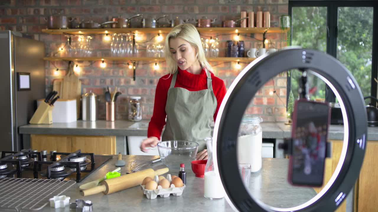 Donna che saluta la telecamera e poi rompe un uovo in una ciotola sull'isola della cucina, mescolando la pastella e mostrando i passaggi