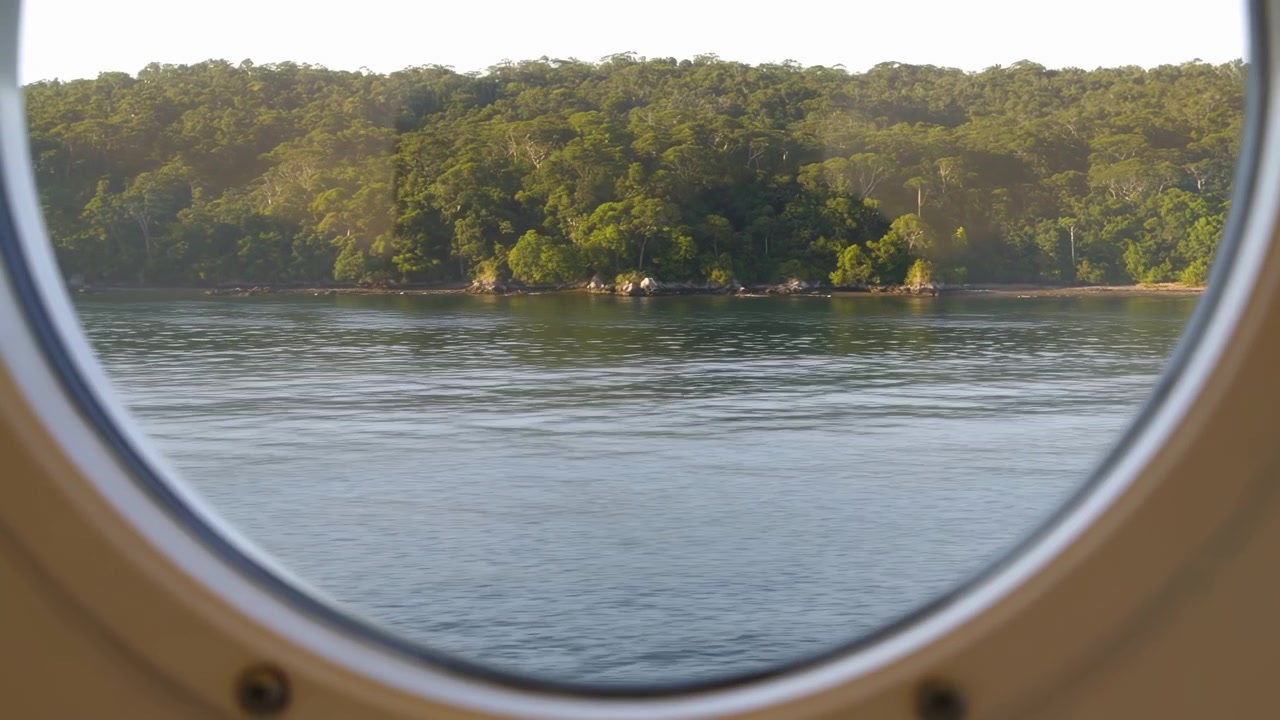 Circular vessel porthole frames calm blue gray water, green rainforest shoreline, and rocky coast under bright hazy daylight, with gentle ripples, soft reflections, and quiet scenic travel mood in
