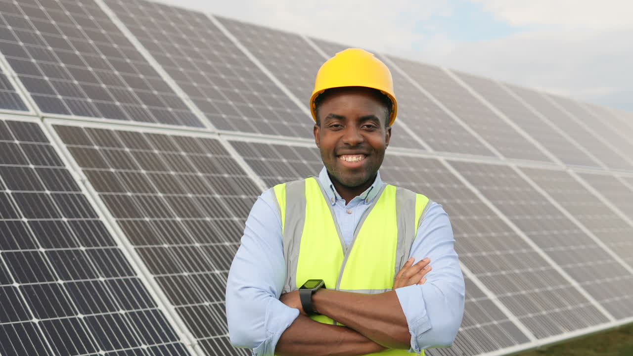 vista de cerca de un joven ingeniero afroamericano con casco protector sonriendo a la cámara con los brazos cruzados en una plantación solar