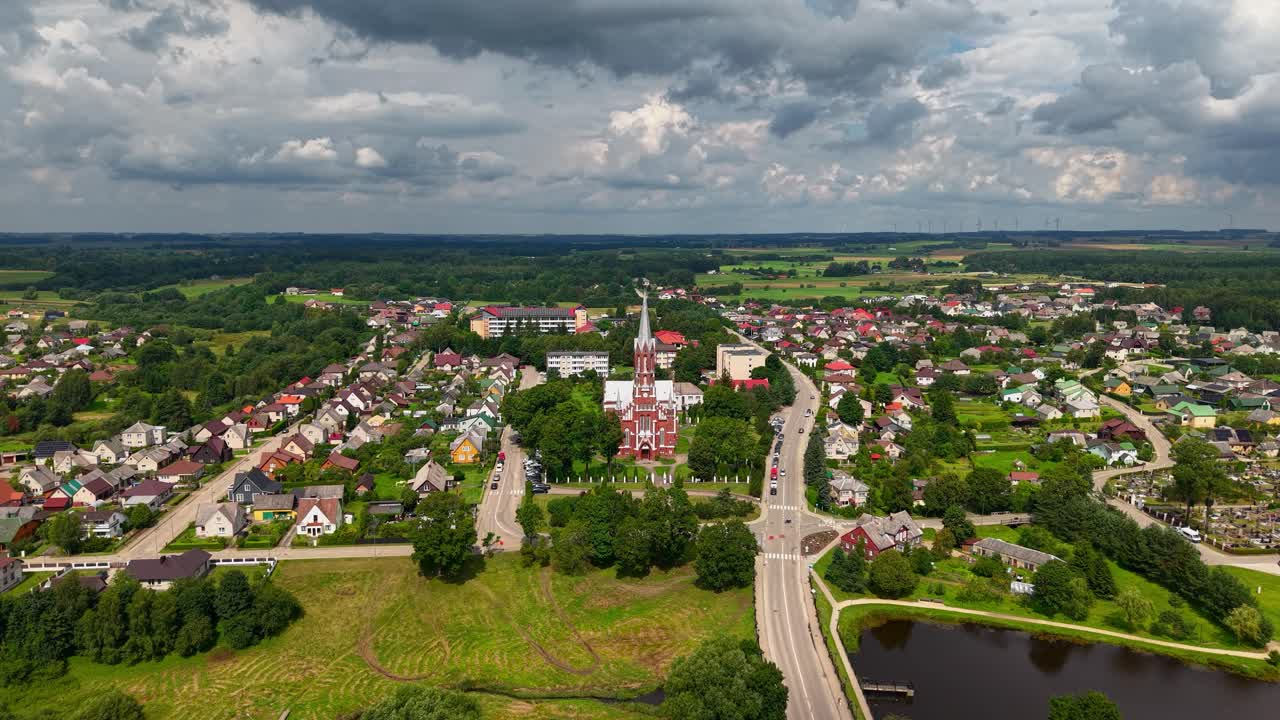 Aerial panoramic view of Šilalė town in Lithuania featuring St. Francis of Assisi Church under dramatic stormy skies, surrounded by colorful houses and lush countryside