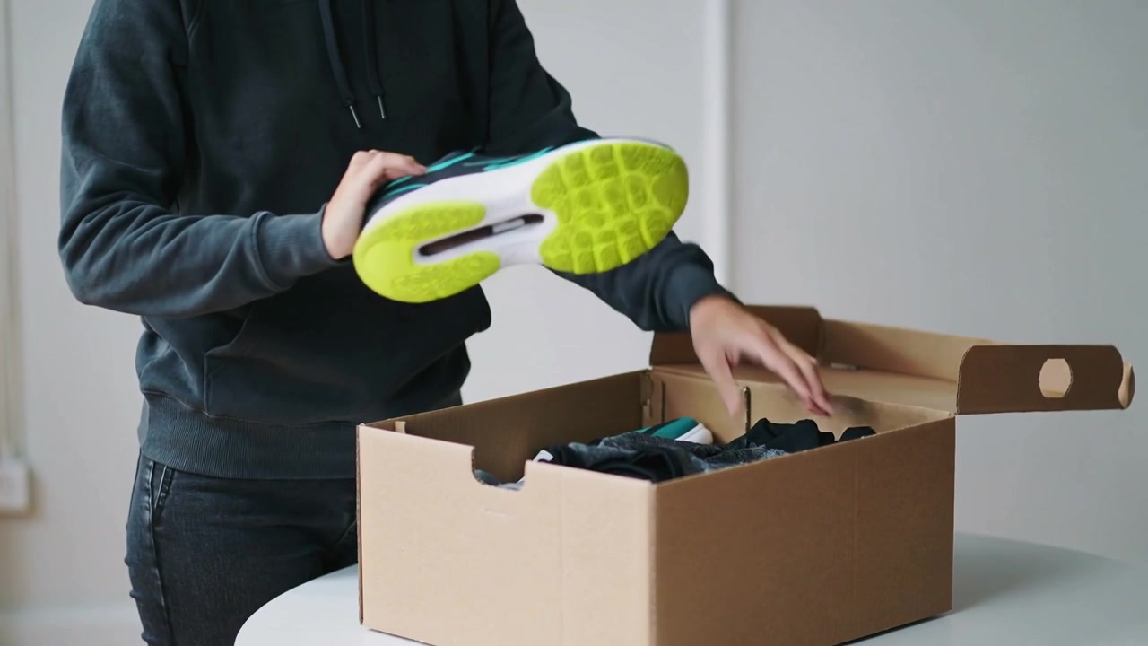 Opening shoebox lid, man in dark hoodie removing sneaker and inspecting shirt at round white table