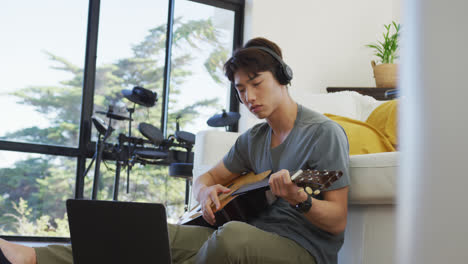 asian boy wearing headphones playing guitar looking at the laptop at home