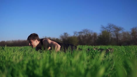 A-fit,-attractive-man-goes-exercising-on-a-green-grass.-Does-push-ups-with-outstretched-hands.-A-beautiful-background.-Black-trousers-and-T-shirt.-Slow-motion