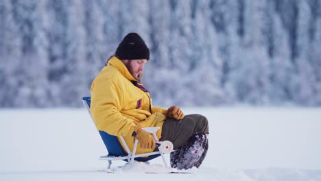 side portrait of a man sitting in portable folding fishing chair, catching fish in ice hole with ice fishing rod in winter
