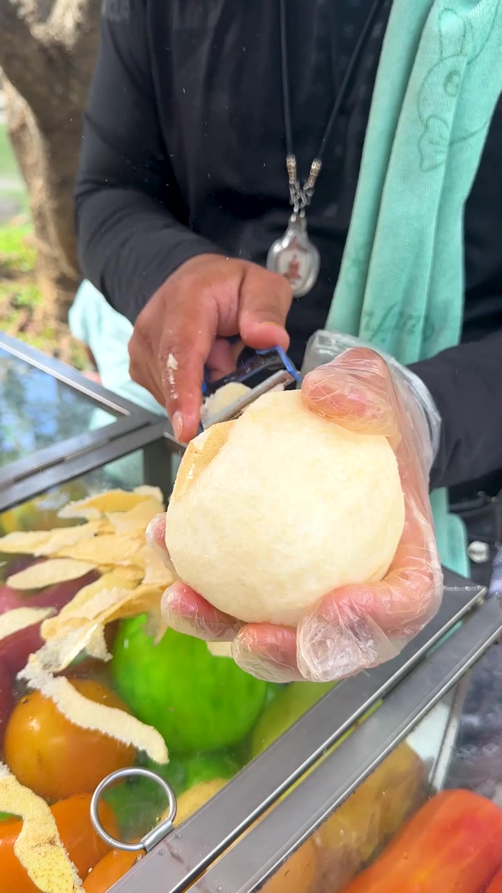 Penjual bersarung tangan mengupas jambu biji dengan pisau di pasar terbuka, di bawah cahaya alami, dari perspektif jarak dekat.