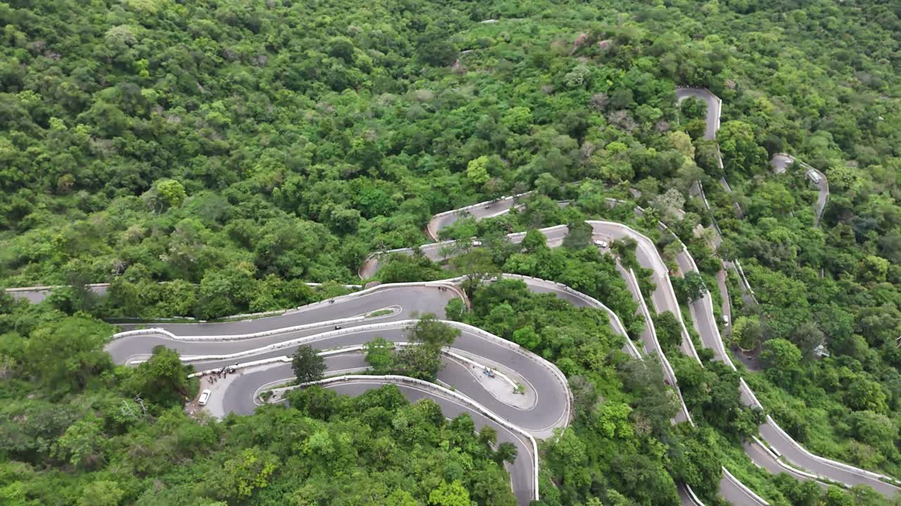 Wide shot showing the early, curvy sections of the Kolli Hills 70-hairpin road. The road crosses farmed red-soil land before entering the dense forest slopes