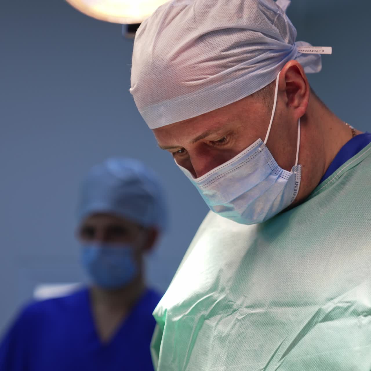 Experienced male surgeon in cap and mask looks down at the patient. Low angle view. Illumination equipment of surgery room at backdrop