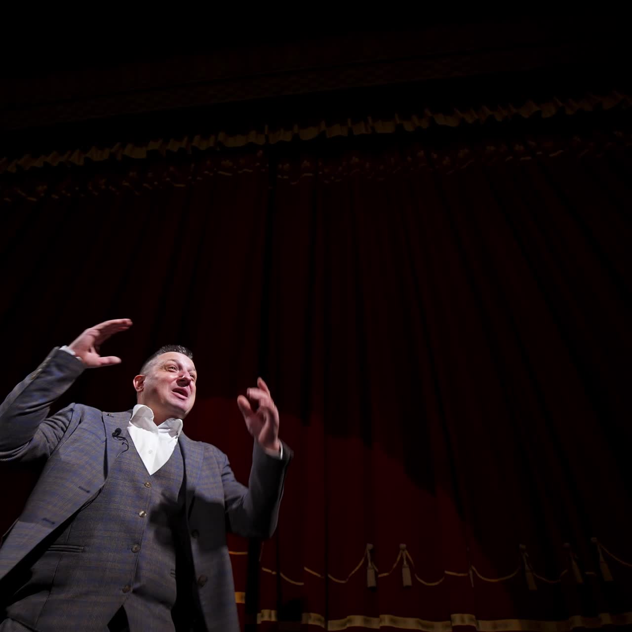 Actor performing on stage. Man in grey suit speaking with many gestures on red theatrical curtains background. View from below