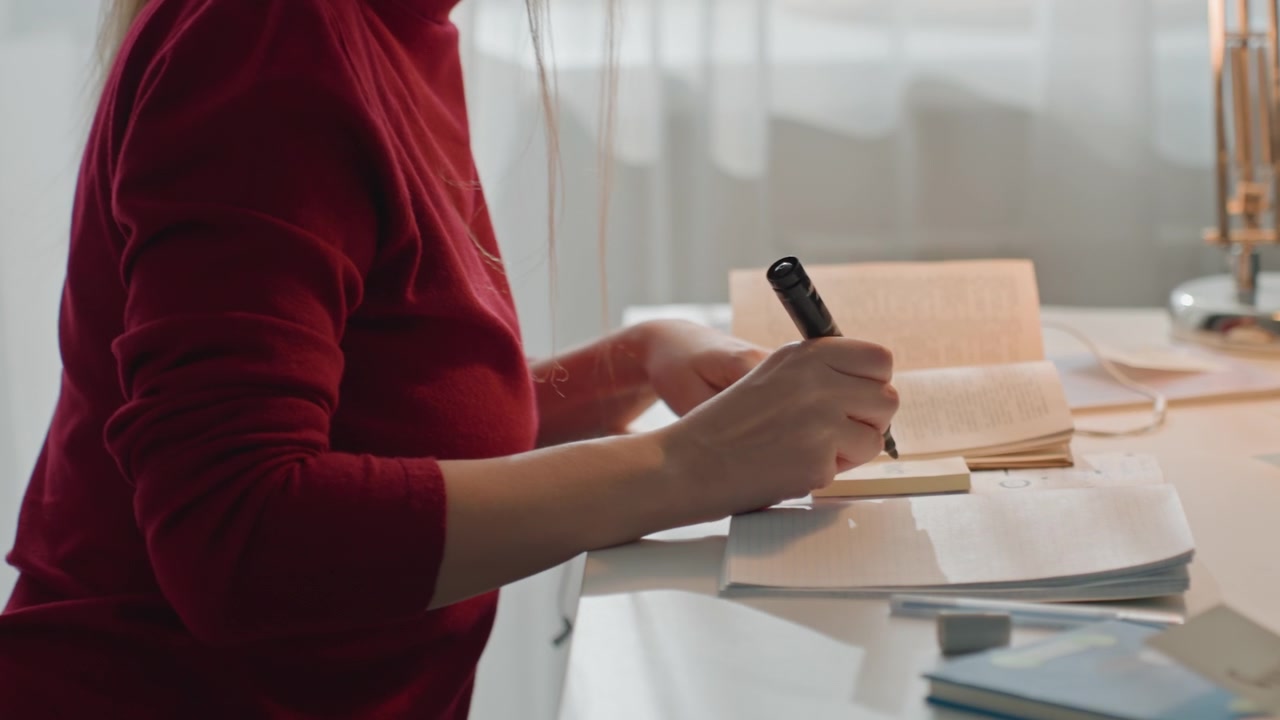 Young woman in red top writes on sticky notes and organizes papers. Woman uses black marker to mark cards labeled. Scene is well lit by natural light through sheer curtains