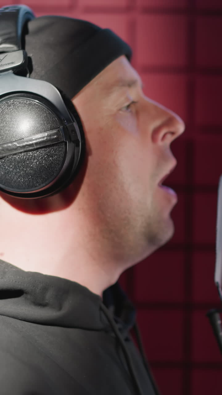 close-up lateral de un hombre vestido de negro, de pie frente a un micrófono de estudio con auriculares, cantando apasionadamente con movimiento de la mano visible en una habitación a prueba de sonido con paneles acústicos rojos