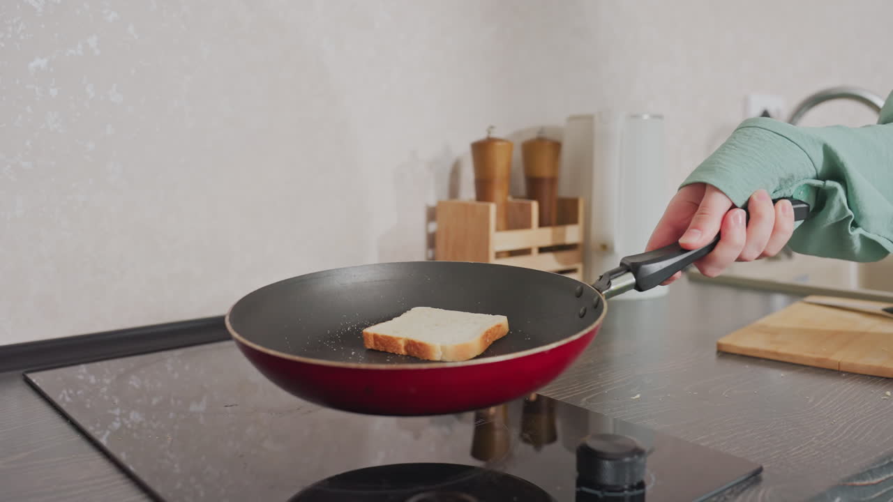 woman in green shirt tossing slice of bread in red frying pan on electric stove while preparing breakfast in bright modern kitchen with cutting board, electric kettle, spice rack on dark countertop
