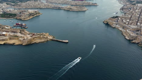 Aerial-view-of-boats-driving-in-front-of-the-cityscape-of-Malta-on-a-cloudy-evening