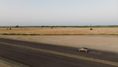 grey car driving on an unfinished tarmac road, accelerating, aerial tracking wide shot