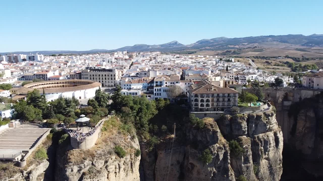 High drone shot captures scenic town with white buildings and historic Puente Nuevo bridge stretching across steep rocky gorge under clear sky, showing dramatic landscape and urban contrast.